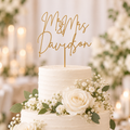 A two-tier white wedding cake decorated with white roses, baby's breath, and green leaves, topped with a gold 'Mr. & Mrs.